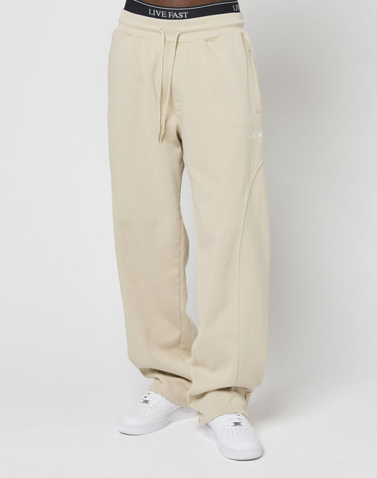 Someone stands against a white background wearing LIVE FAST DIE YOUNG’s Basic Wide Jogger in beige with an elastic waistband and wide fit, paired with white sneakers and black underwear featuring LIVE FAST on the waistband.