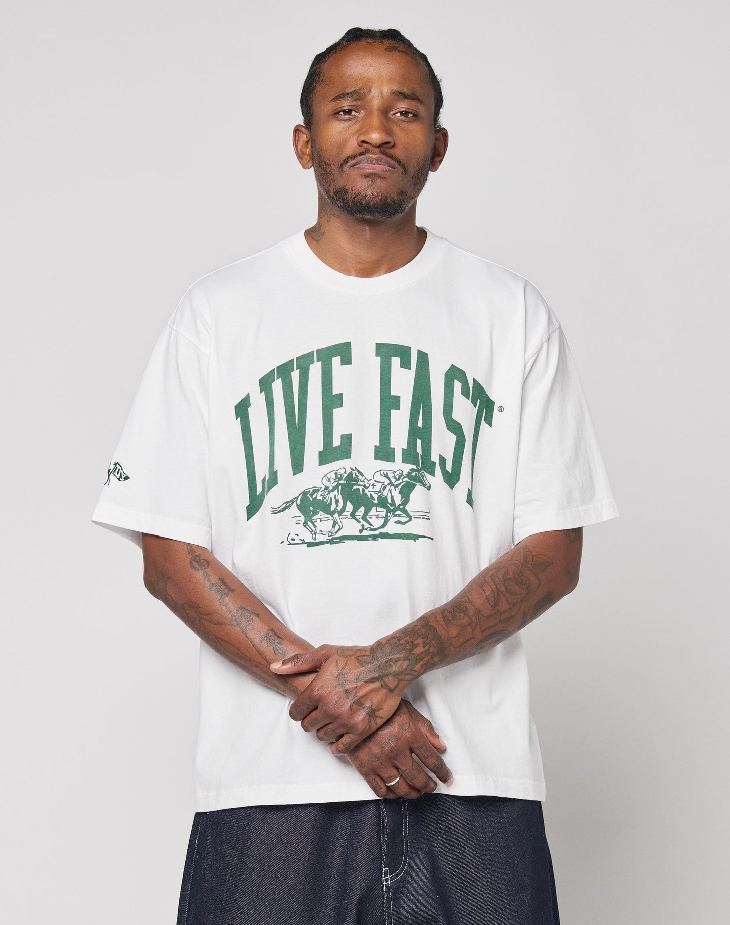 A tattooed man faces the camera wearing the Live Fast Stallion Tee by LIVE FAST DIE YOUNG, an oversize white 100% Baumwolle T-shirt with a green LIVE FAST and running horses print, paired with dark pants against a plain light background.