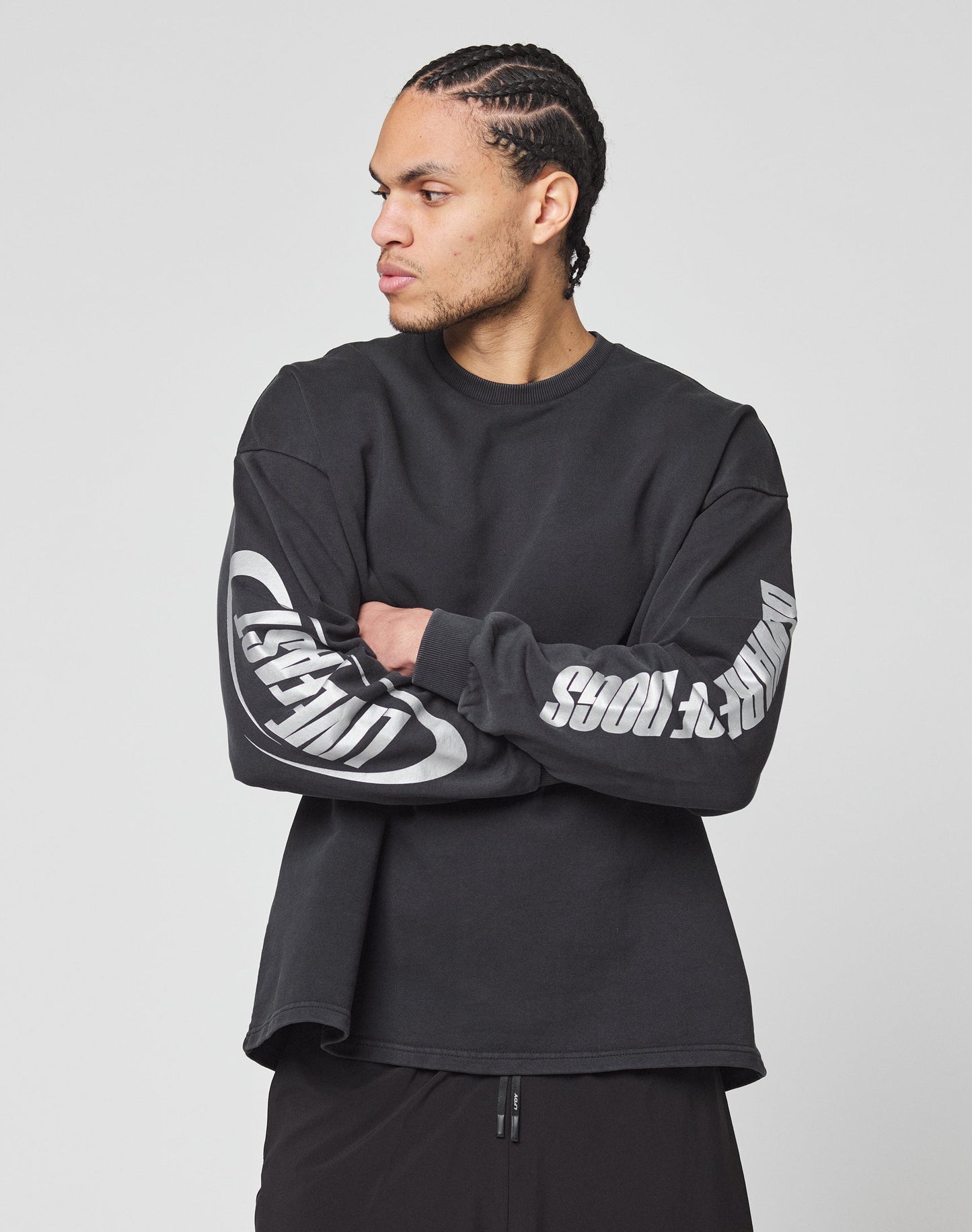 A man with braided hair wears the LFDY Canine Longsleeve by LIVE FAST DIE YOUNG—black, 100% Baumwolle, with bold white reflektierender Druck and sleeve graphics. He stands arms crossed, facing left against a plain light background.