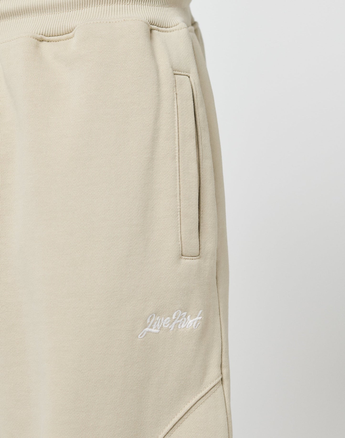 Close-up of Basic Wide Jogger sweatpants by LIVE FAST DIE YOUNG in beige, featuring a wide fit, side pockets, elastic waistband, and white Live Fast embroidery near the hip. Made from soft cotton. Plain light background.