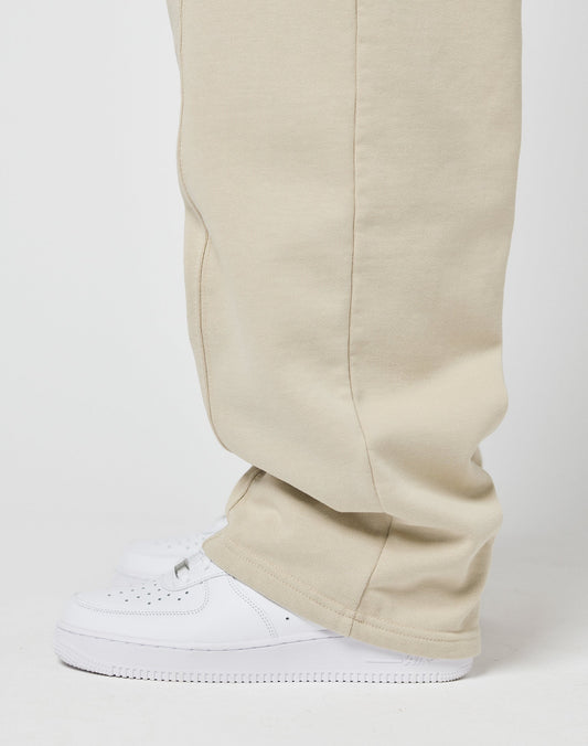 Side view of a person in beige LIVE FAST DIE YOUNG Basic Wide Jogger pants with elastic waistband, paired with white sneakers. The lower legs and feet are visible, pants slightly covering the shoes against a plain background.