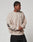 A person wears the LIVE FAST DIE YOUNG Live For It Sweater, a loose beige sweatshirt with black text on the left sleeve and chest, standing against a plain background and looking to the side with hands loosely clasped in front.