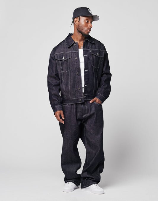 A man in an LFDY Denim Jacket by LIVE FAST DIE YOUNG, matching baggy jeans, a white T-shirt, white sneakers, and a dark baseball cap stands against a plain light background, looking to his left with a relaxed posture.