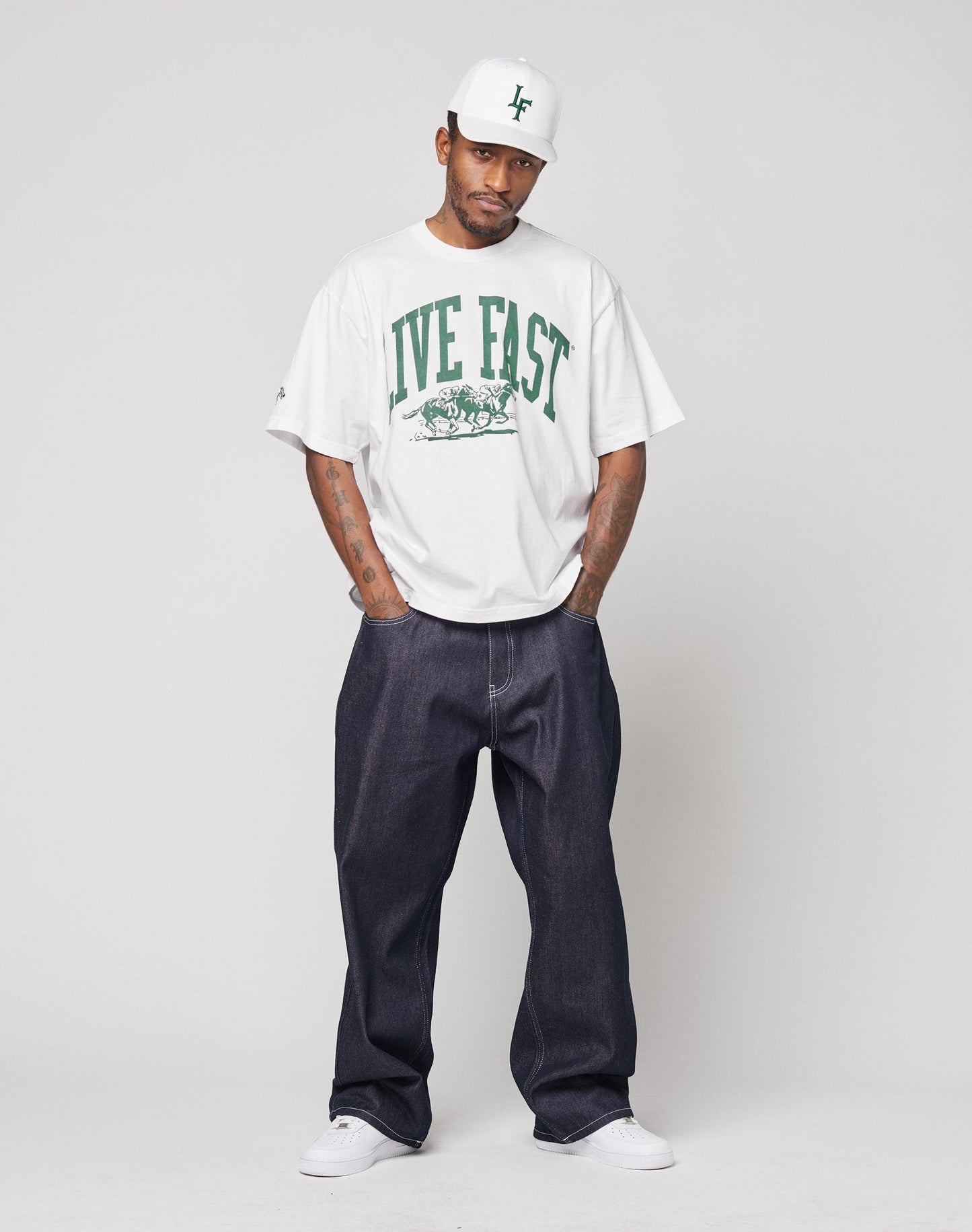 A man wears a white oversize Live Fast Stallion Tee by LIVE FAST DIE YOUNG, paired with a white cap, dark baggy jeans, and white sneakers, standing with hands in pockets against a plain light background.