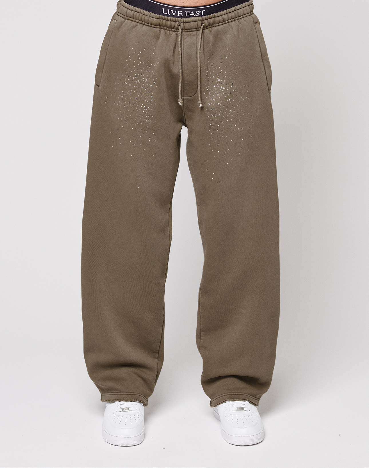 A person wears the Live Fast Rhinestone Jogger by LIVE FAST DIE YOUNG—olive green, drawstring waist, rhinestones on the upper front, and an oversized fit—styled with white sneakers and black LIVE FAST underwear peeking above the waistband.