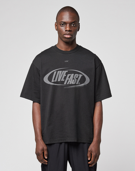 A person wearing the LFDY Sphere Tee by LIVE FAST DIE YOUNG—an oversized black cotton T-shirt with "LIVE FAST." boldly printed on the front—stands against a plain, light gray background. | model