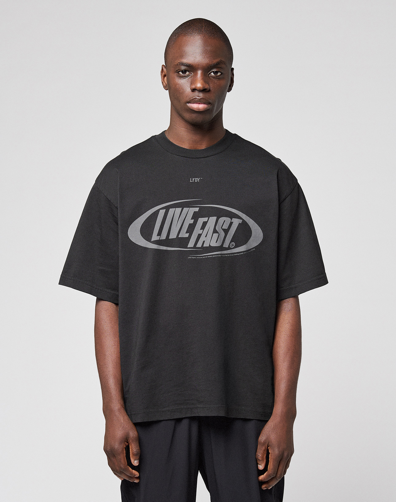 A person wearing the LFDY Sphere Tee by LIVE FAST DIE YOUNG—an oversized black cotton T-shirt with "LIVE FAST." boldly printed on the front—stands against a plain, light gray background. | model