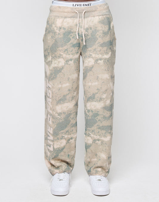 A person wearing LIVE FAST DIE YOUNG’s Live Fast Boucle Pants in light green and beige tie-dye with a broad elastic waistband, white drawstrings, and ‘LIVE FAST’ vertically on one leg, paired with white sneakers against a plain background.