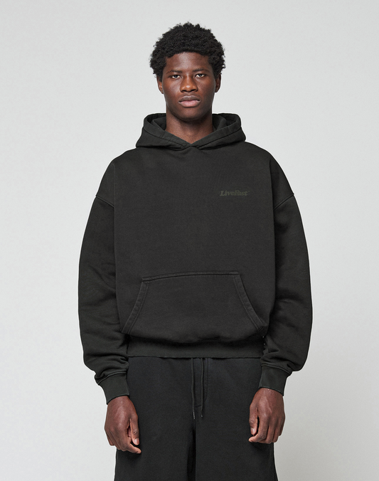 A young man with short, curly hair faces forward, wearing the LIVE FAST DIE YOUNG Basic 360 Hooded in black with front and back print and black pants against a plain light gray background.