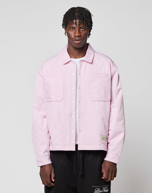 A man with short, braided hair wears the LIVE FAST DIE YOUNG LFDY Striped Padded Shirt—an oversized, light pink and white striped design with large front pockets—over a white shirt and black pants, standing against a plain light gray background.