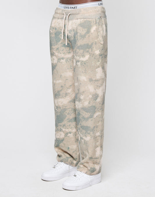 A person wears LIVE FAST DIE YOUNG's Live Fast Boucle Pants in relaxed beige and light blue tie-dye with a wide elastic waistband, white drawstring, and visible LIVE FAST branding—styled with white sneakers for a comfy, oversized look. | model