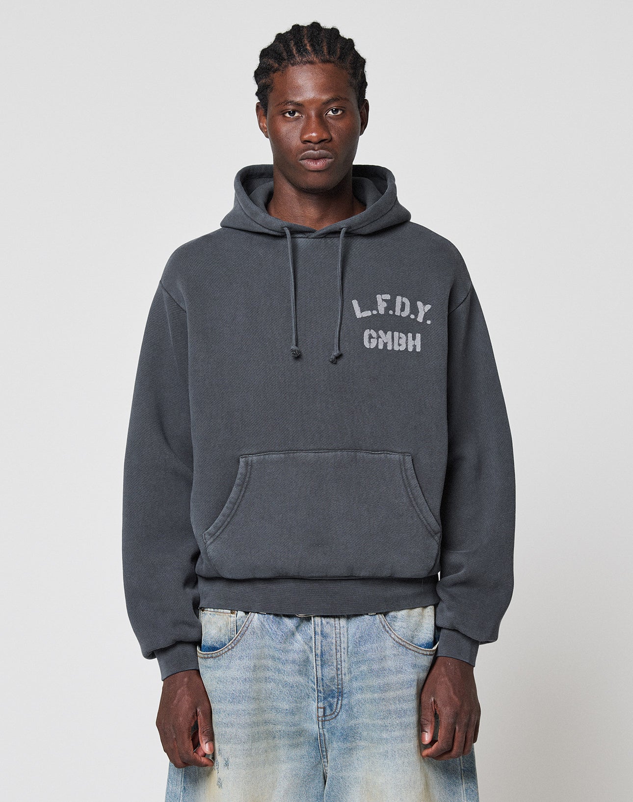 A man wears the LIVE FAST DIE YOUNG LFDY GmbH Hooded in dark gray with “L.F.D.Y. GMBH” on the chest and an oversize fit, styled with light blue faded jeans against a plain light background. | model