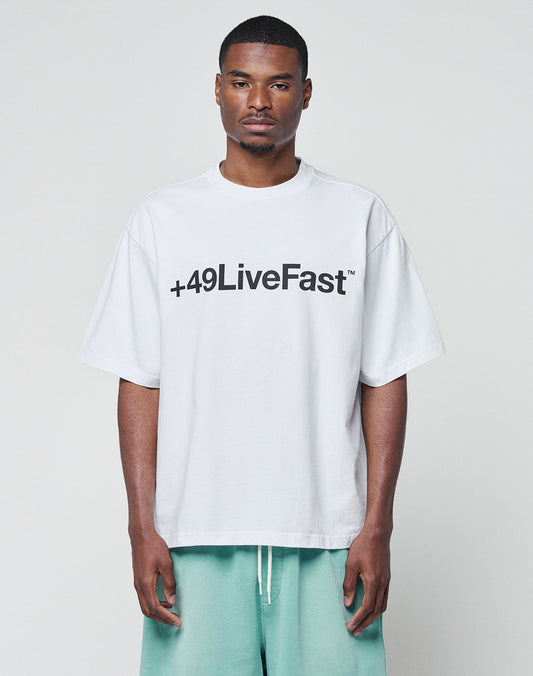 A man wears the LIVE FAST DIE YOUNG Basic +49 Tee, an oversize fit white T-shirt with a bold black +49LiveFast front print, made from 100% Baumwolle. He pairs it with light green shorts and stands against a light gray background.