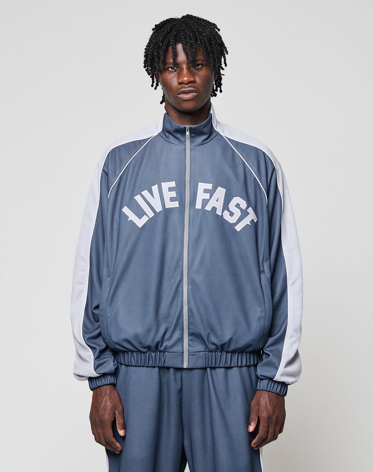 A man with medium-length curly hair wears a blue and gray LFDY Arch Trackjacket by LIVE FAST DIE YOUNG, featuring LIVE FAST printed in white on the front. He stands facing forward with hands in the side pockets against a plain light background. | model