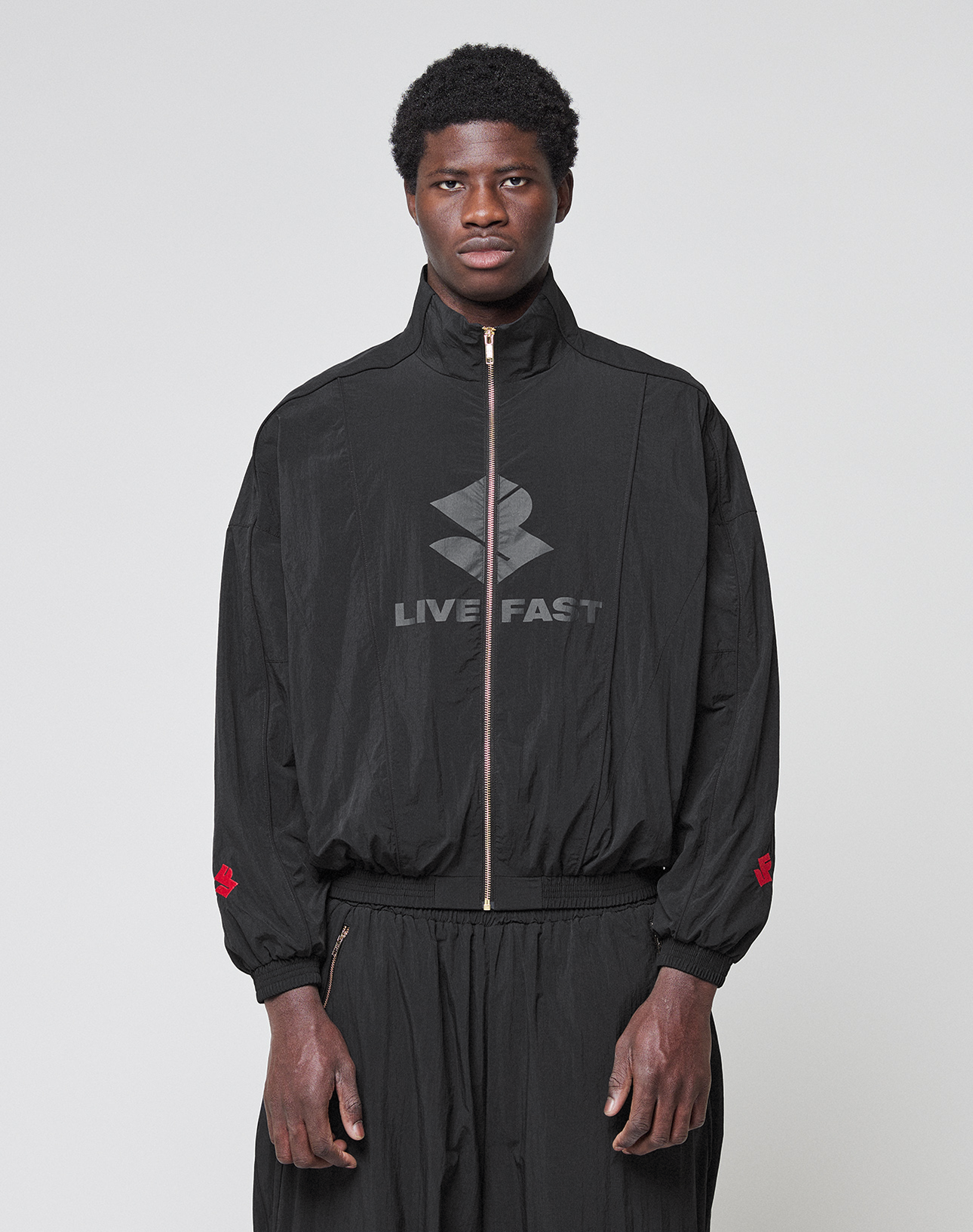 A man stands facing forward, wearing the LFDY Dealership Trackjacket by LIVE FAST DIE YOUNG—black with a stand-up collar, zip-up front with gold zipper, Live Fast logo, and red sleeve accents—against a plain light background.