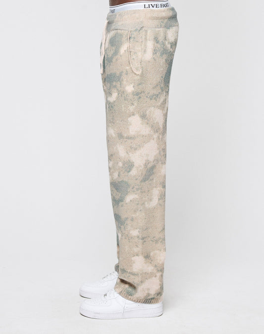 A person in Live Fast Boucle Pants by LIVE FAST DIE YOUNG, featuring tan and green tie-dye, a breiter Elastikbund, wide fit, and gerades Bein with "LIVE FAST" on the waistband, stands in profile with white sneakers against a white background.