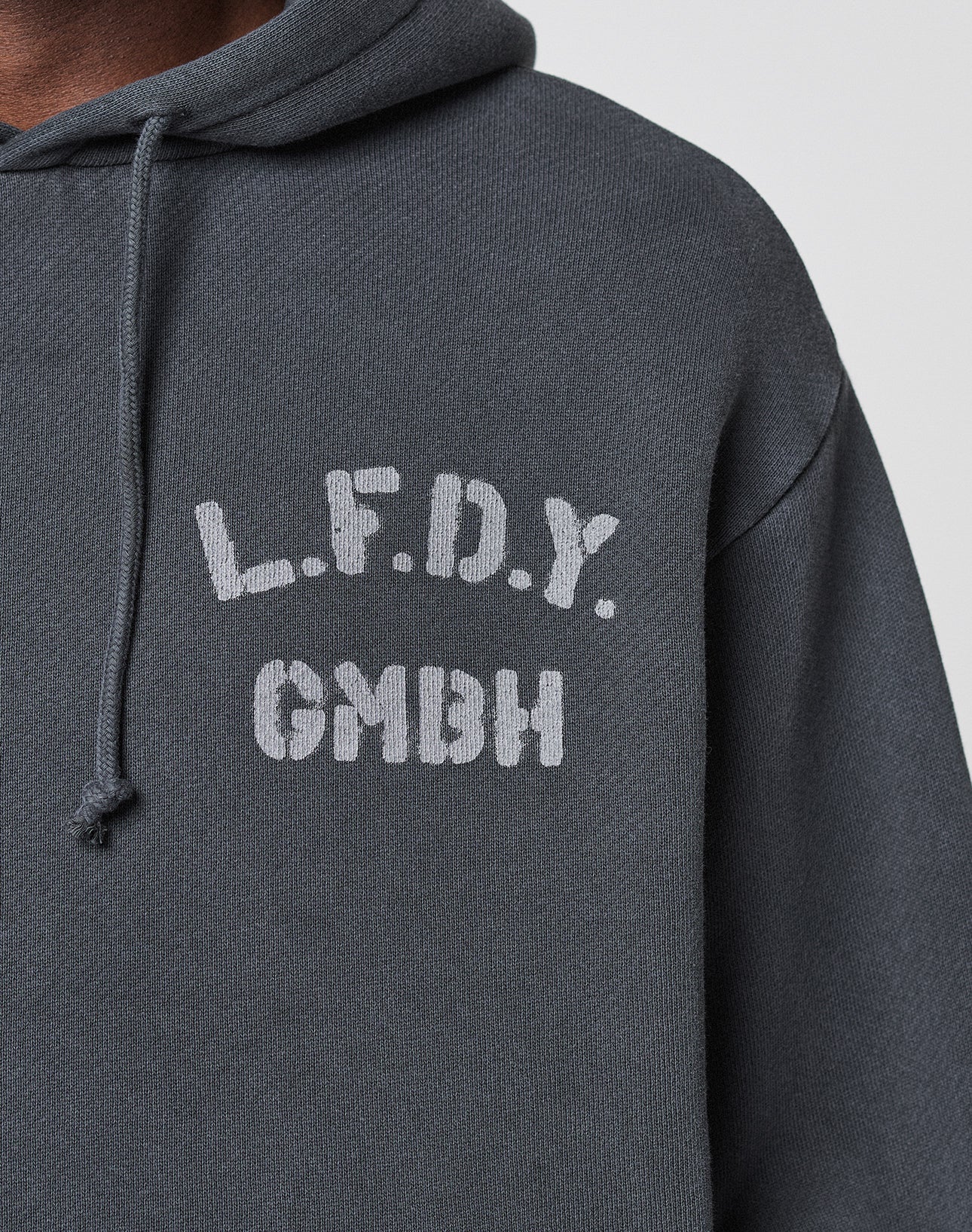 A close-up of the LFDY GmbH Hooded by LIVE FAST DIE YOUNG, a dark gray oversized hoodie with white "L.F.D.Y. above GMBH" text on the left chest and a visible drawstring on the left side of the hood.