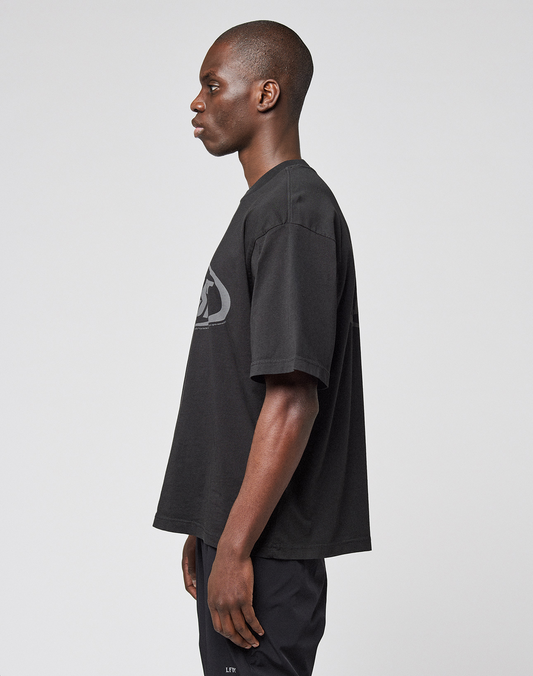 A person stands in profile against a plain background, wearing the LFDY Sphere Tee by LIVE FAST DIE YOUNG—an oversized black Baumwolle T-shirt with subtle front and back prints—paired with black pants. Their arms rest relaxed at their sides.