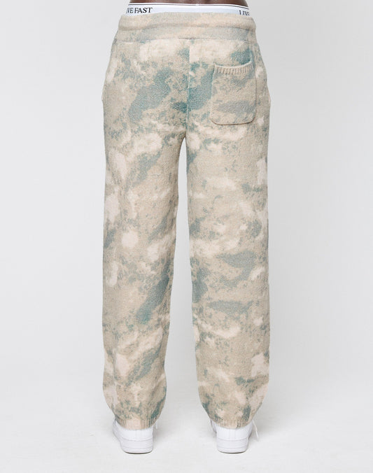 A person seen from the back wears LIVE FAST DIE YOUNG’s Live Fast Boucle Pants in beige and green tie-dye with a wide elastic waistband, straight leg, single rear pocket, and white shoes, set against a plain white background.