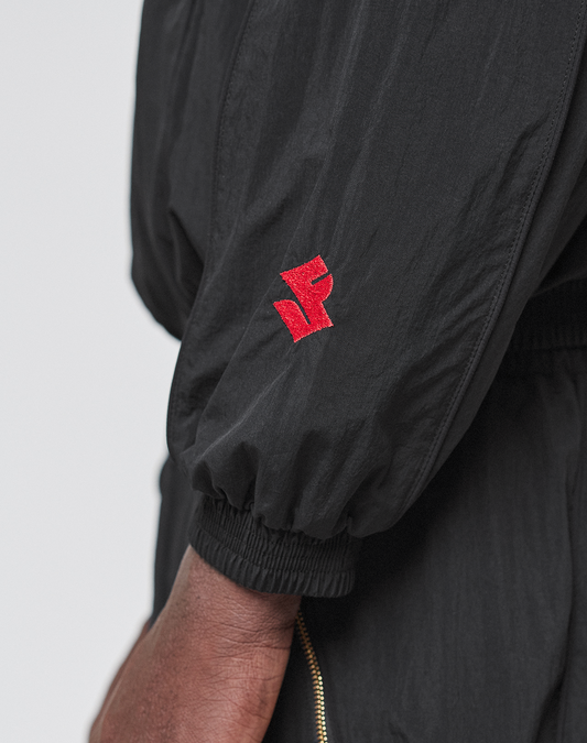 A close-up of an arm in the LIVE FAST DIE YOUNG LFDY Dealership Trackjacket, featuring a red embroidered logo on the sleeve, regular fit, elastic cuffs, stand-up collar (Stehkragen), and a visible gold zipper.