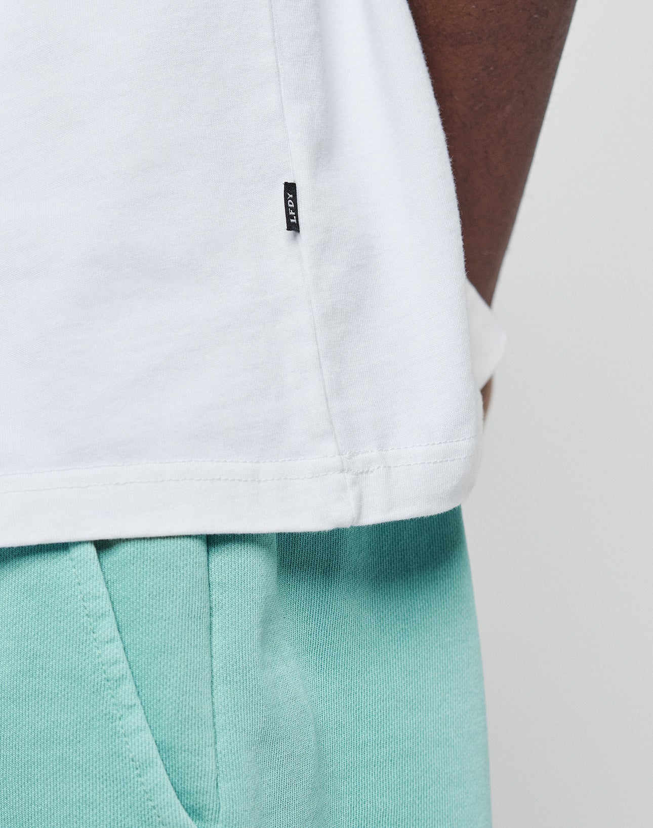 Close-up of a person wearing an oversize fit white Basic +49 Tee by LIVE FAST DIE YOUNG, made from 100% Baumwolle, and mint green sweatpants. Only part of the torso and hand are visible. A small black tag is on the T-shirt.