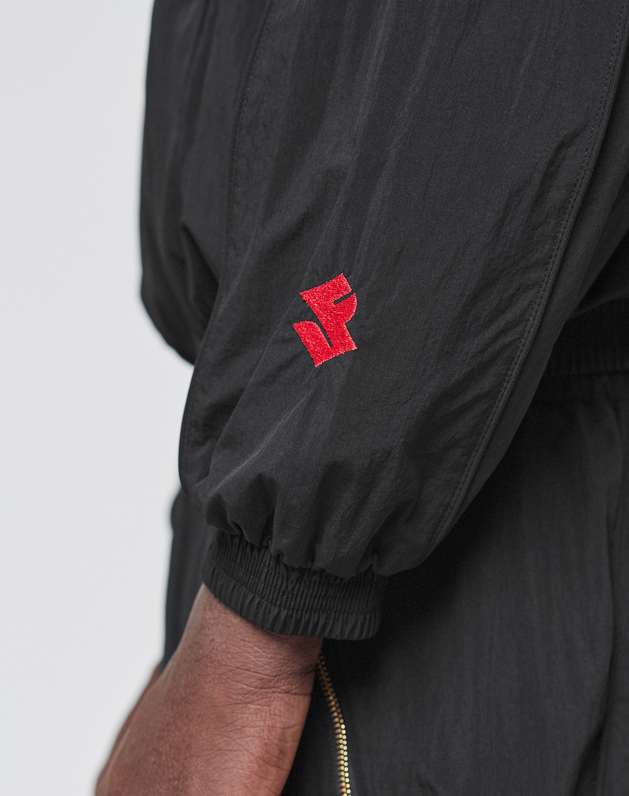A close-up of an arm in the LIVE FAST DIE YOUNG LFDY Dealership Trackjacket, featuring a red embroidered logo on the sleeve, regular fit, elastic cuffs, stand-up collar (Stehkragen), and a visible gold zipper.