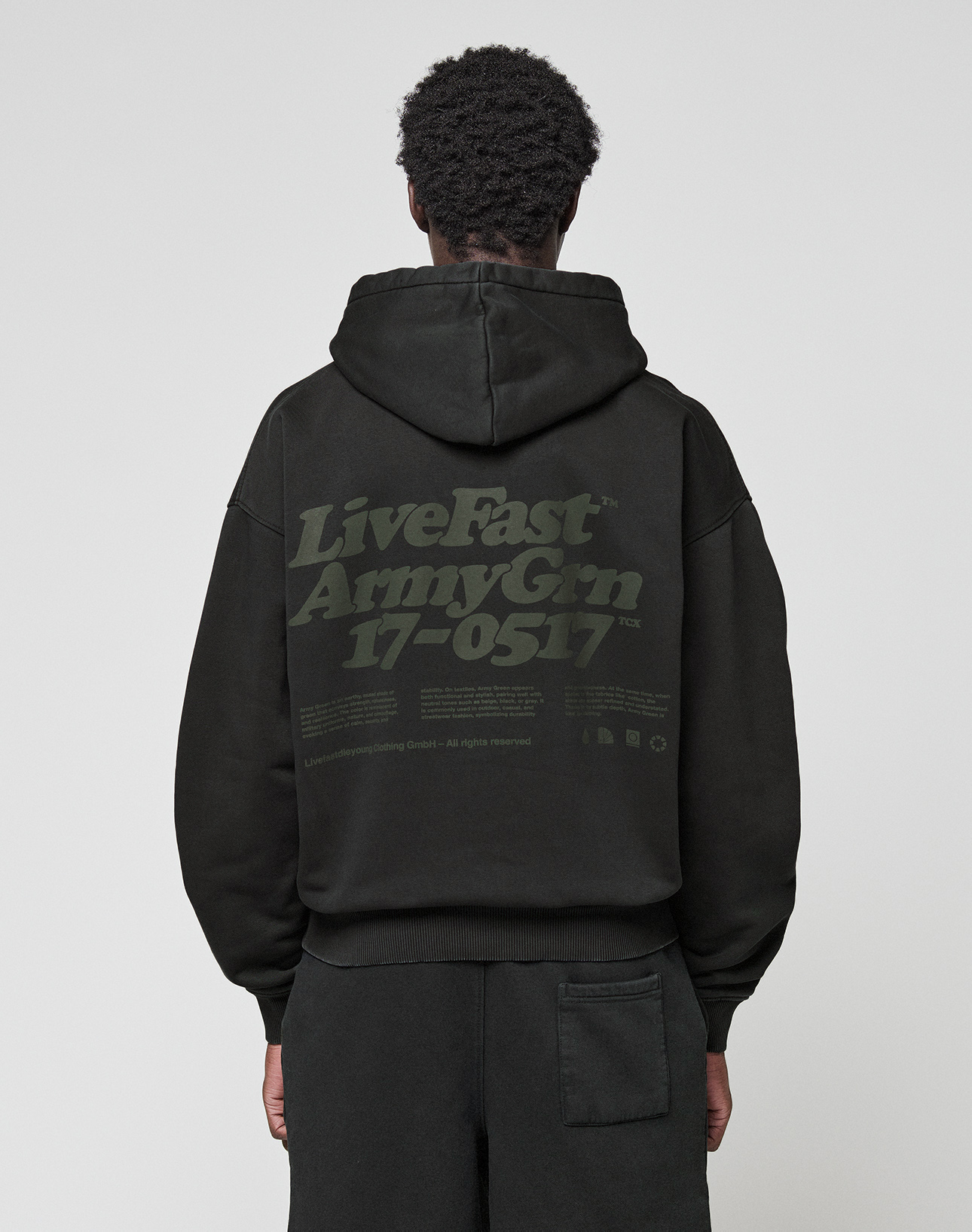 A person wearing the LIVE FAST DIE YOUNG Basic 360 Hooded black hoodie with the hood up, seen from behind. The back showcases large green "LiveFast ArmyGrn 17-0517" text and small writing—a stylish front and back print design. | model