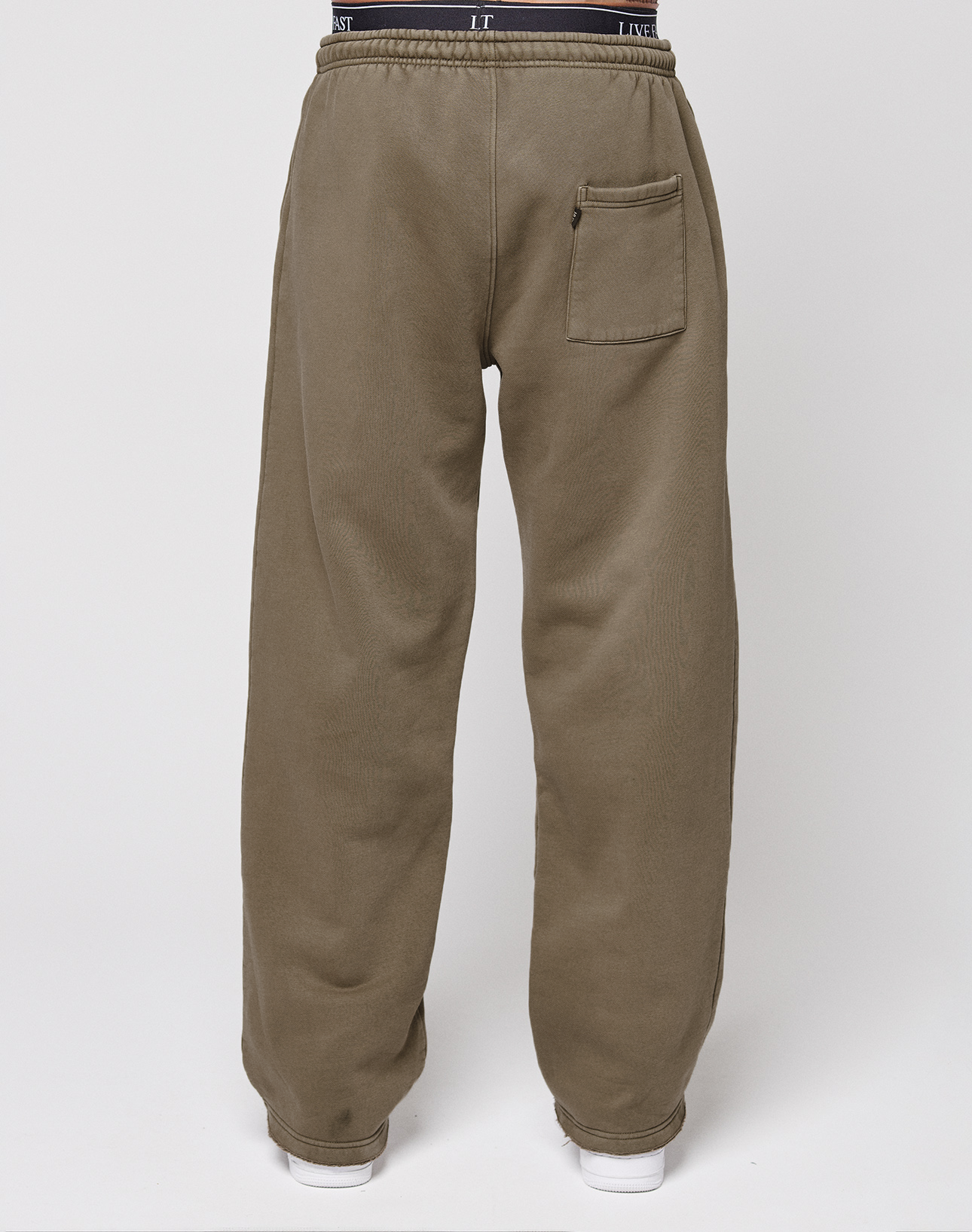 A person wears the LIVE FAST DIE YOUNG Live Fast Rhinestone Jogger, featuring loose olive fabric, an oversize fit, elastic waistband, and single back pocket, standing against a plain white background.