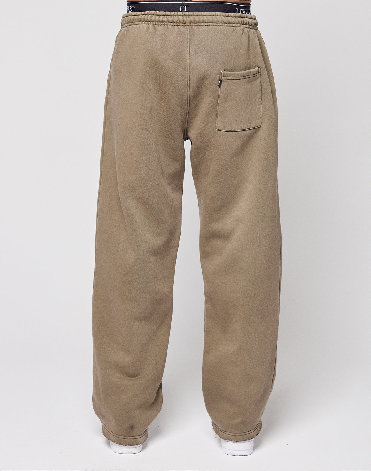 A person stands with their back to the camera, wearing LIVE FAST DIE YOUNG's Live Fast Rhinestone Jogger—light brown, loose-fit sweatpants with a vintage wash, elastic waistband, and single back pocket—against a plain white background.