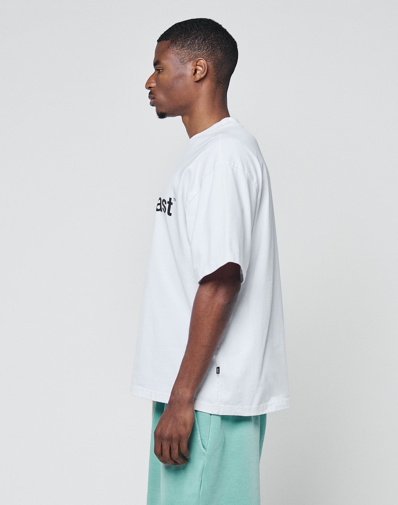 A man in profile poses against a plain background, wearing a LIVE FAST DIE YOUNG Basic +49 Tee in white with partial black text and light green shorts, both made from 100% Baumwolle.