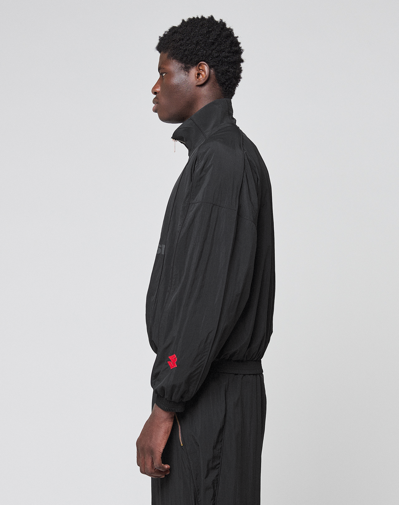A person with short curly hair stands in profile against a plain background, wearing the LIVE FAST DIE YOUNG LFDY Dealership Trackjacket in black with a Stehkragen, matching black pants, and a small red logo on the sleeve.