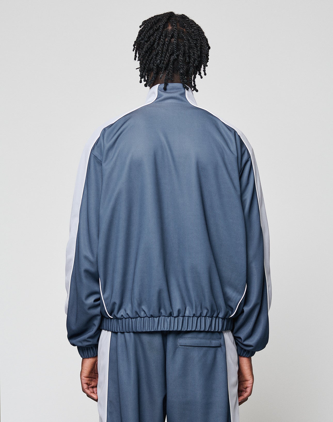 A person with short twists is seen from behind, wearing the LIVE FAST DIE YOUNG LFDY Arch Trackjacket in blue with white side stripes, Oversize Fit, high collar, and elastic cuffs against a plain light background.