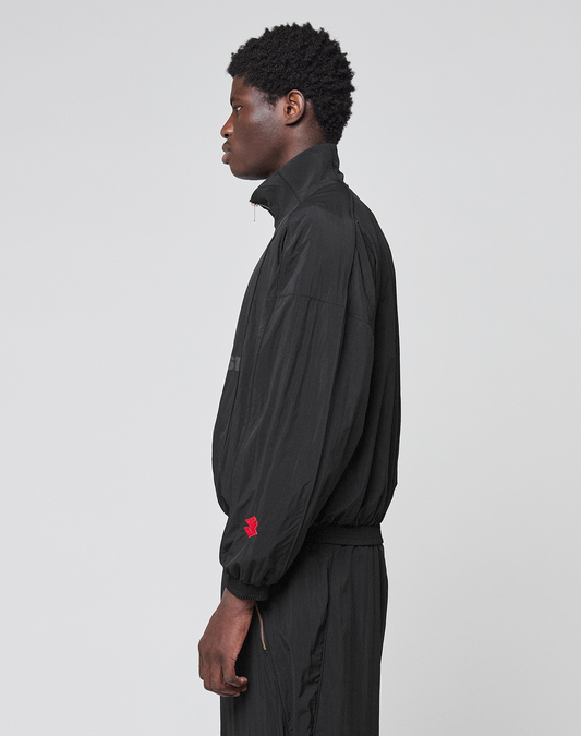 A person with short curly hair stands in profile against a plain background, wearing the LIVE FAST DIE YOUNG LFDY Dealership Trackjacket in black with a Stehkragen, matching black pants, and a small red logo on the sleeve.
