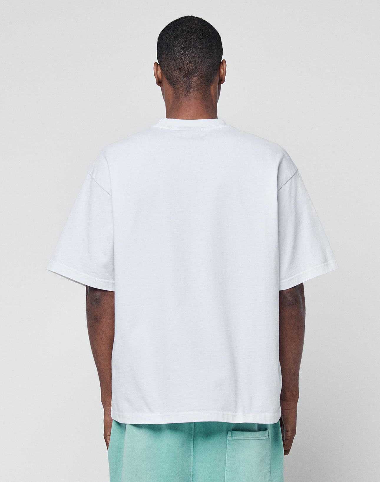 A person faces away from the camera, wearing the LIVE FAST DIE YOUNG Basic +49 Tee in white and mint green shorts, set against a plain light gray background.
