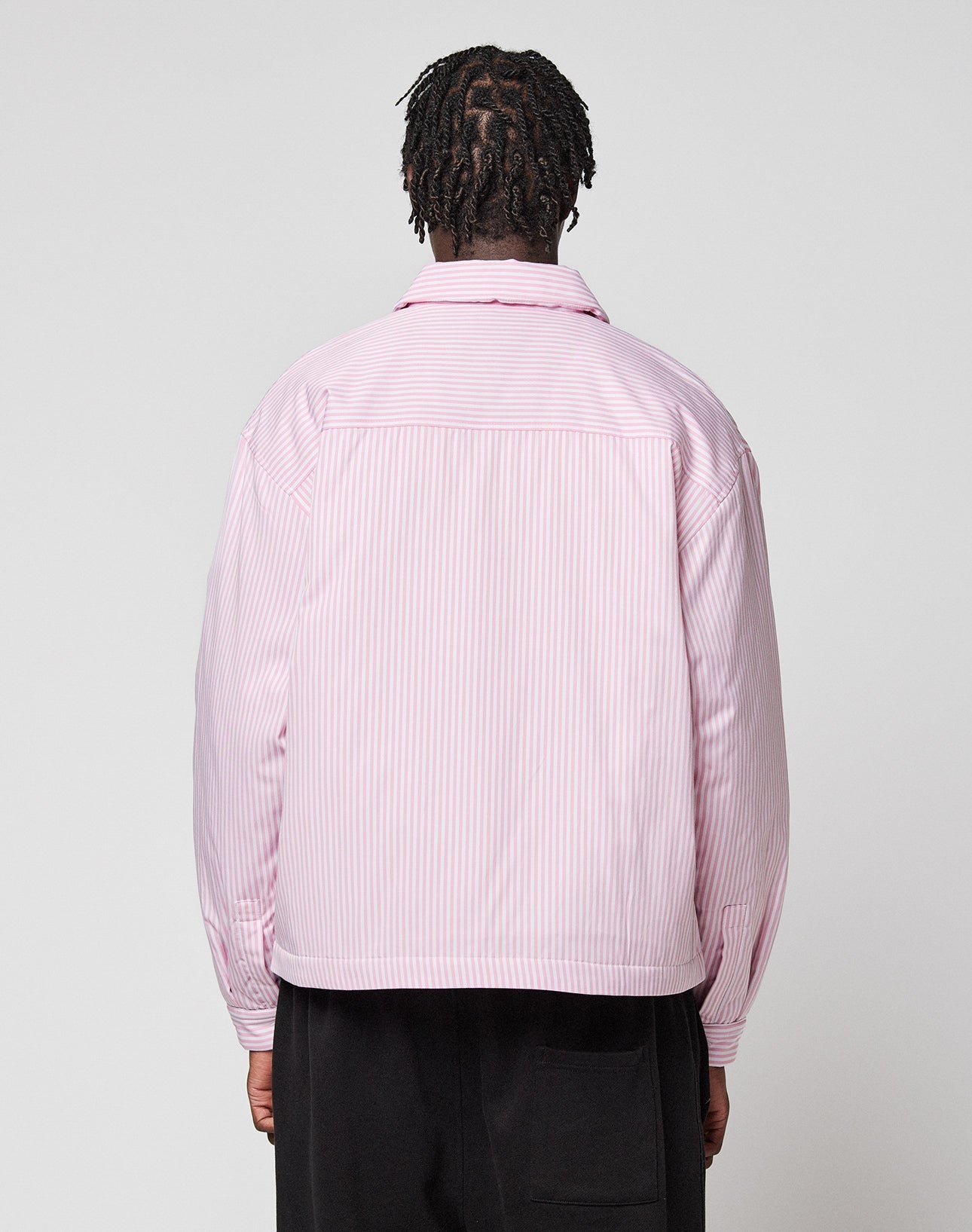 A person with short dreadlocks faces away, wearing the LIVE FAST DIE YOUNG LFDY Striped Padded Shirt in light pink and white stripes and loose-fitting black pants, standing against a plain light gray background.