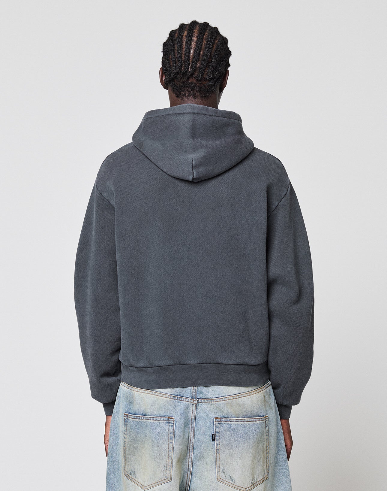 A person with braided hair is shown from the back in a dark gray LFDY GmbH Hooded by LIVE FAST DIE YOUNG and light blue jeans, set against a plain, light background.