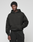 A young man with short curly hair wears the LIVE FAST DIE YOUNG Basic 360 Hooded black hoodie and matching pants with front and back print, standing against a plain light gray background, relaxed with one hand in his pocket.