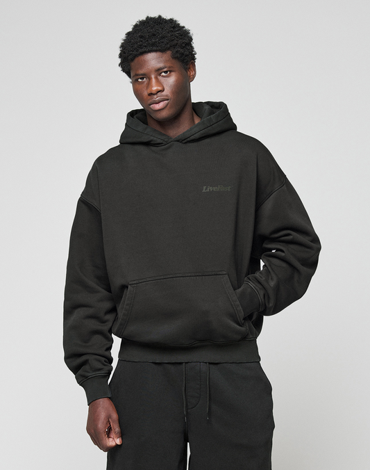 A young man with short curly hair wears the LIVE FAST DIE YOUNG Basic 360 Hooded black hoodie and matching pants with front and back print, standing against a plain light gray background, relaxed with one hand in his pocket.