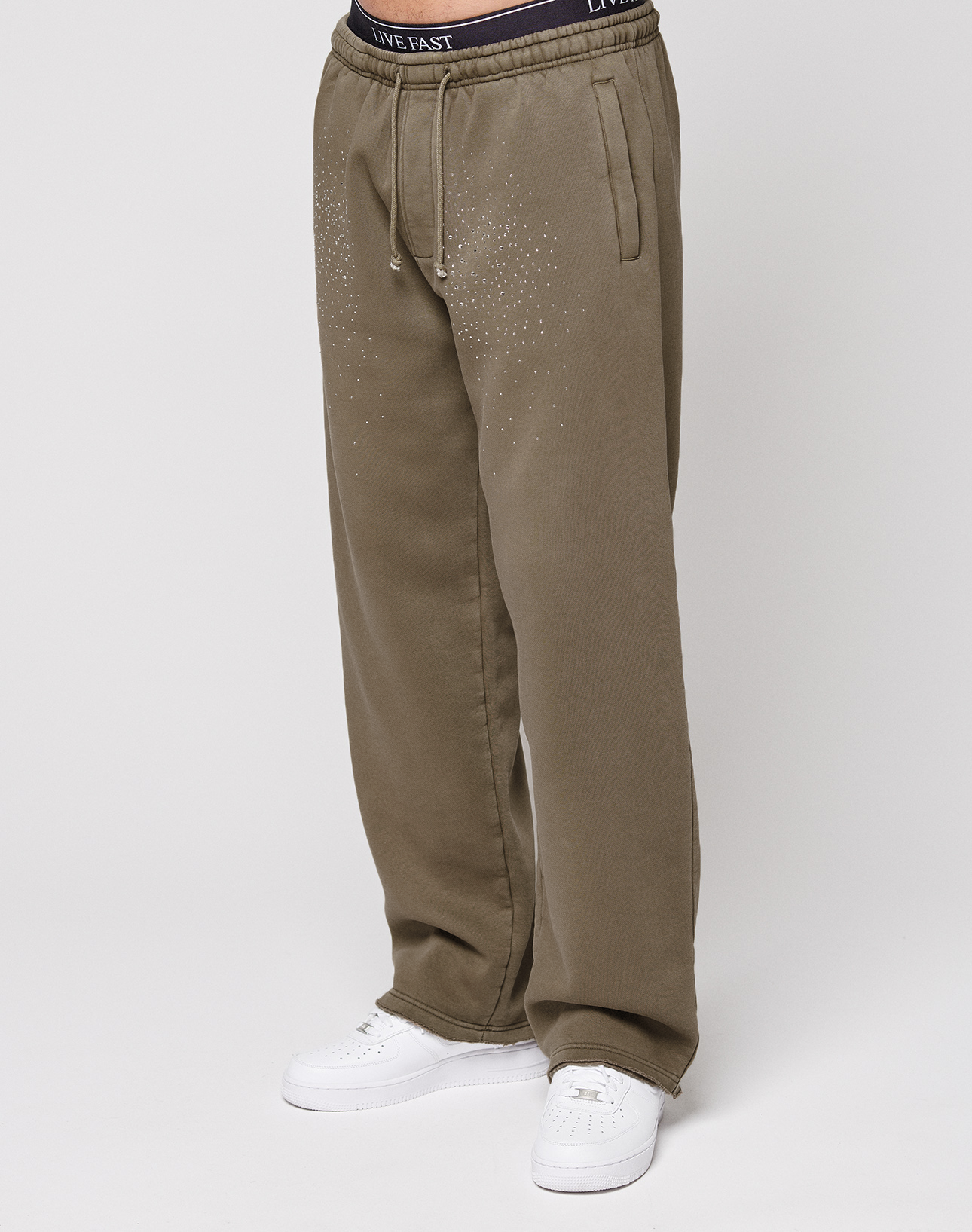 A person wears the LIVE FAST DIE YOUNG Live Fast Rhinestone Jogger—loose-fitting olive green sweatpants with a vintage wash, rhinestone dot accents on the upper thighs, and a "LIVE FAST" waistband. Paired with white sneakers against a plain white background. | model