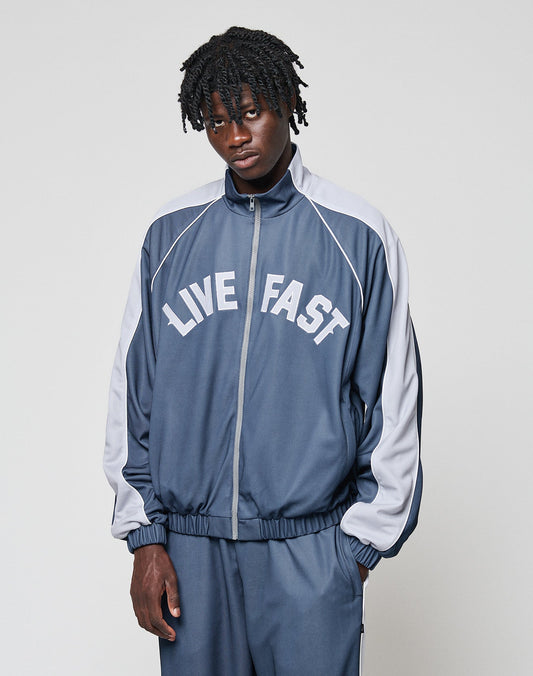 A person with short dreadlocks wears the LFDY Arch Trackjacket by LIVE FAST DIE YOUNG—a blue and gray jacket with LIVE FAST in bold white letters, elastische Bündchen, and Reißverschlusstaschen—against a plain light background.