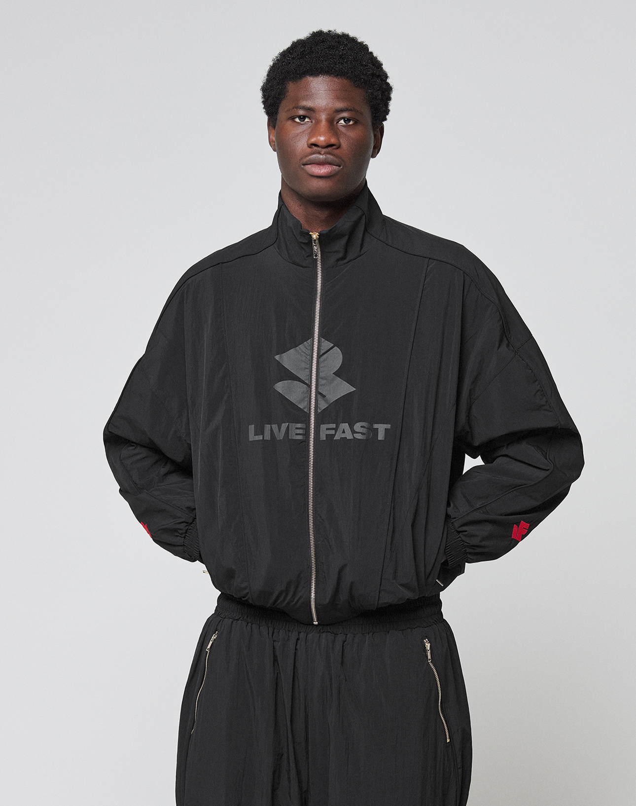 A person wearing the black LFDY Dealership Trackjacket by LIVE FAST DIE YOUNG, featuring a Stehkragen, "LIVE FAST," and a stylized logo on the front, stands against a light gray background with hands in pockets.