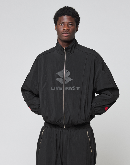 A person wearing the black LFDY Dealership Trackjacket by LIVE FAST DIE YOUNG, featuring a Stehkragen, "LIVE FAST," and a stylized logo on the front, stands against a light gray background with hands in pockets.