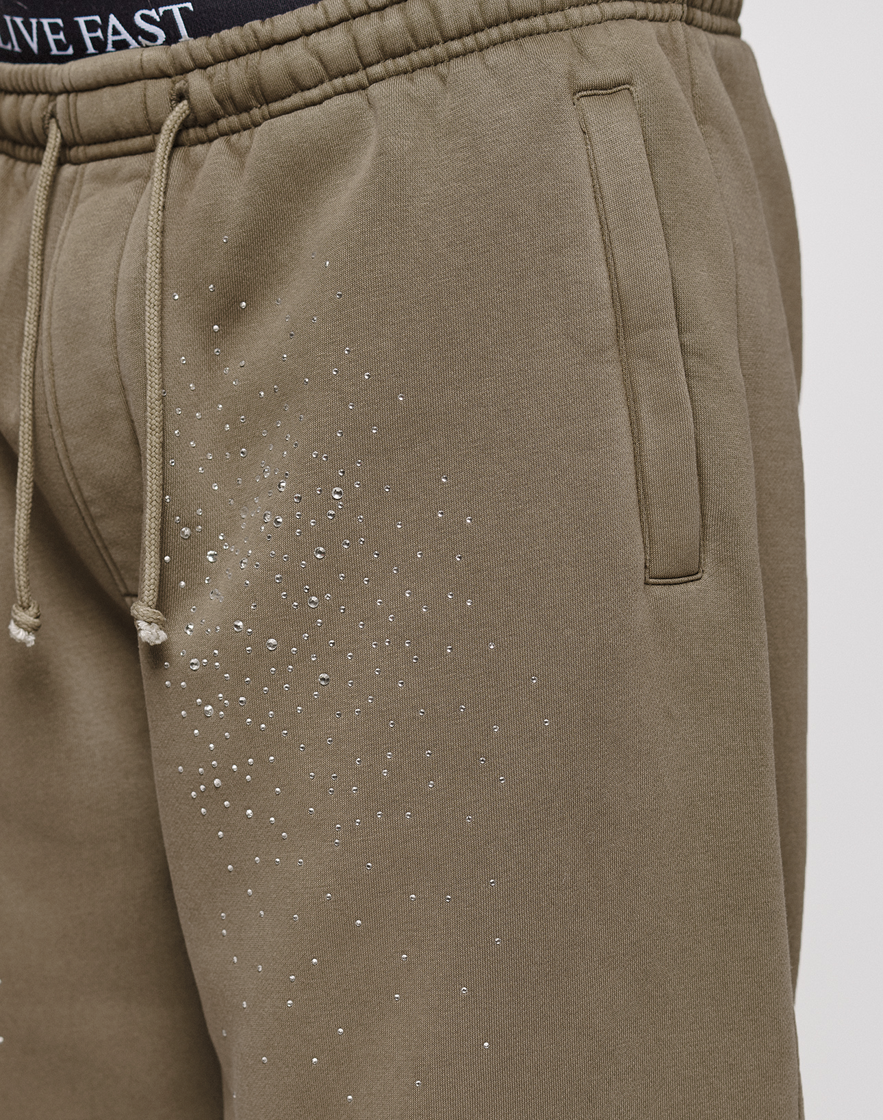 Close-up of a person wearing the LIVE FAST DIE YOUNG Live Fast Rhinestone Jogger in light brown with vintage wash, rhinestone-studded upper thighs, drawstrings, and side pockets. Black LIVE FAST waistband underwear adds an edgy touch.