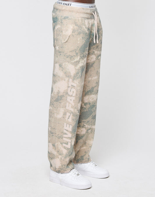 A person wears LIVE FAST DIE YOUNG’s Live Fast Boucle Pants in beige and green tie-dye, featuring a wide elastic waistband, straight leg, and LIVE FAST printed vertically on the left leg, styled with white sneakers against a plain background.