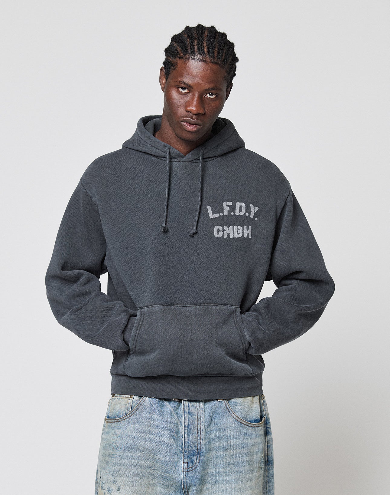A man in a dark grey LIVE FAST DIE YOUNG LFDY GmbH Hooded with an oversize fit, front pocket, and chest print stands against a light background, hands in the pocket, wearing light blue jeans.