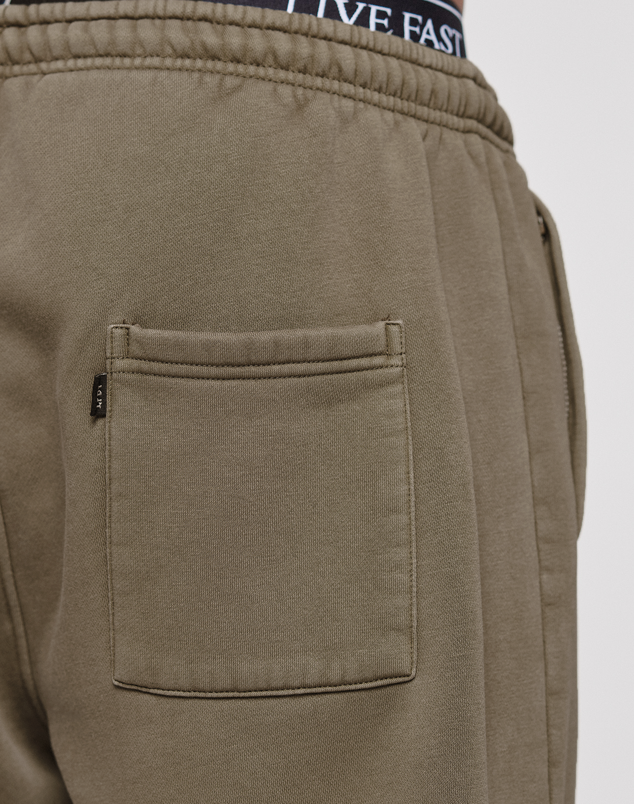 Close-up of someone wearing the Live Fast Rhinestone Jogger by LIVE FAST DIE YOUNG in olive green, showcasing its oversize fit, elastic waistband, and large back pocket. Dark underwear with white lettering peeks above the jogger’s waistband.
