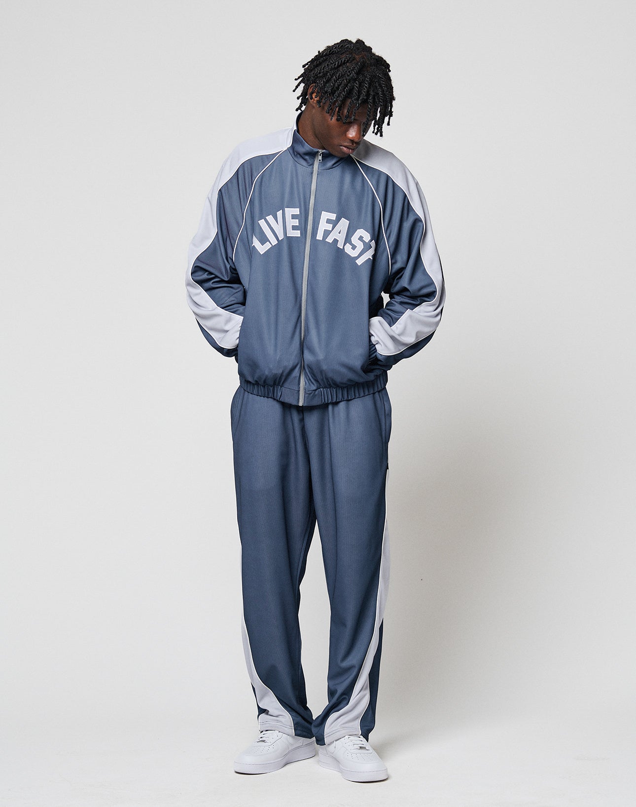 A person stands against a plain background wearing the LFDY Arch Trackjacket by LIVE FAST DIE YOUNG in blue and gray, with LIVE FAST on the jacket, elastic cuffs, zip pockets, hands in pockets, looking down, paired with white sneakers.