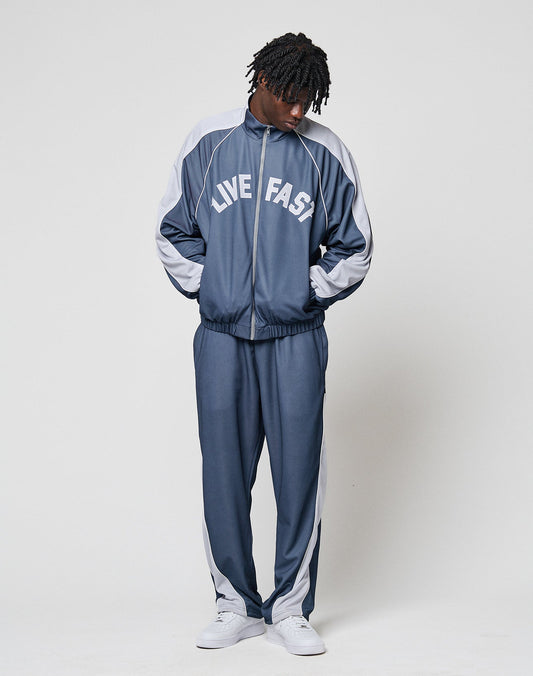 A person stands against a plain background wearing the LFDY Arch Trackjacket by LIVE FAST DIE YOUNG in blue and gray, with LIVE FAST on the jacket, elastic cuffs, zip pockets, hands in pockets, looking down, paired with white sneakers.