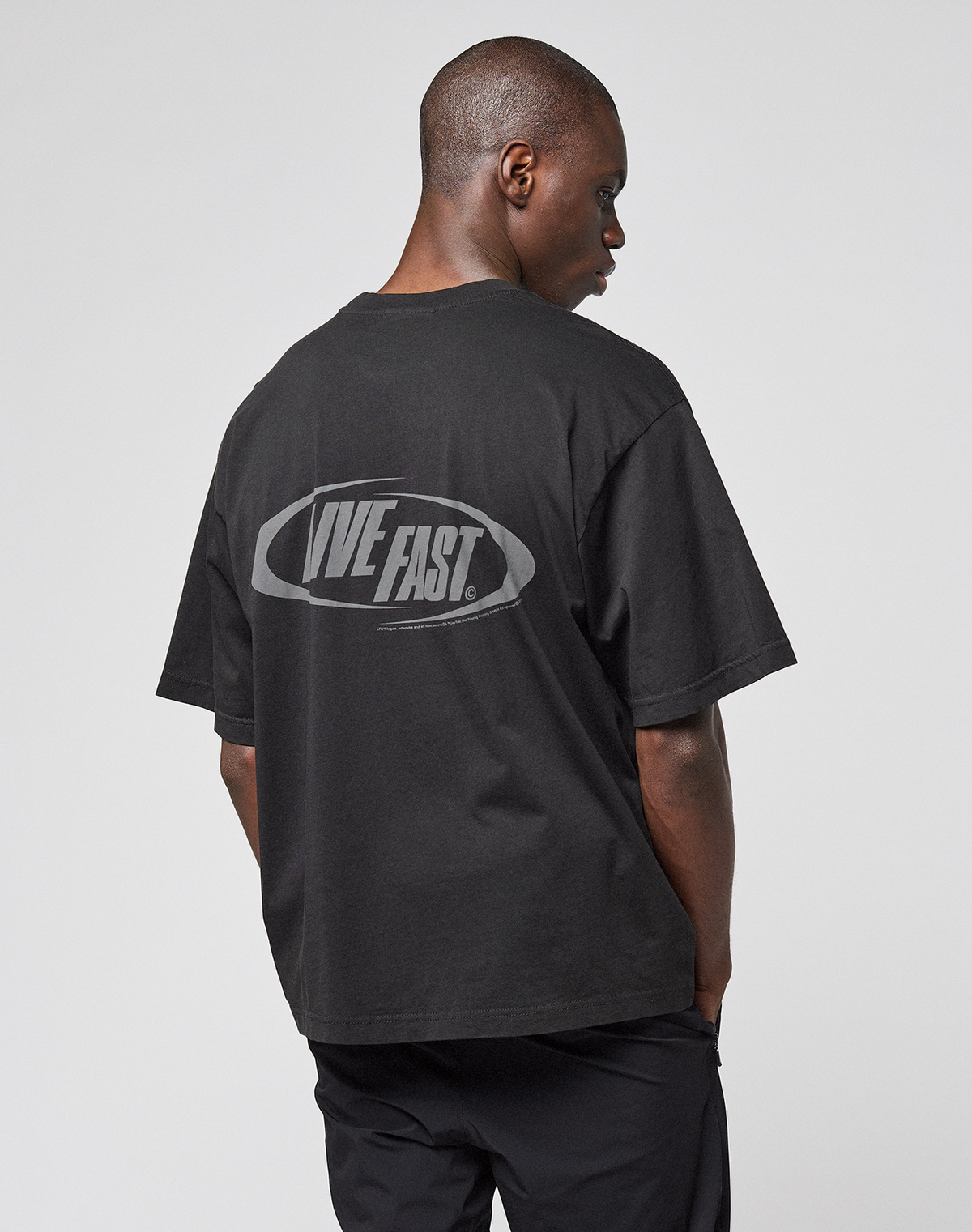 A person faces away, wearing the LIVE FAST DIE YOUNG LFDY Sphere Tee—a black, oversized cotton t-shirt with a bold "LIVE FAST" backprint. The background is plain and light-colored.