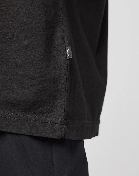 Close-up of the lower side of an oversize black LFDY Sphere Tee by LIVE FAST DIE YOUNG, featuring a small HEY tag near the hem, styled with dark pants. Displayed against a plain, light background.